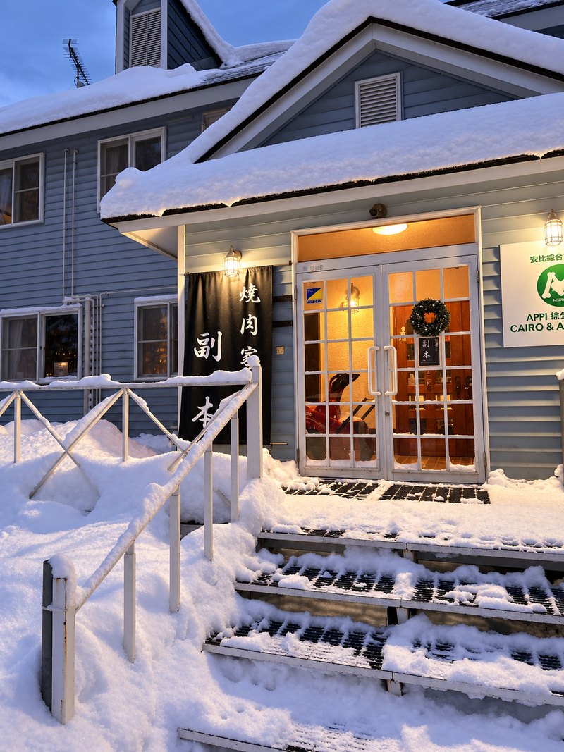 雪の積もった階段と手すりの先に、暖色の灯りがともる店舗の入口と、店名を記した紺色の幟